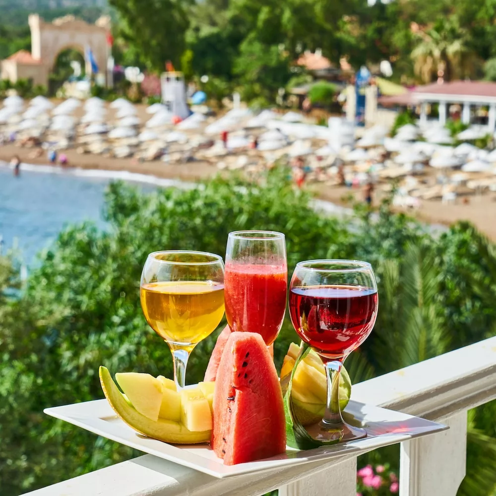 Plate of summer fruit and drinks on a balcony overlooking a beach.